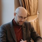 Bald man with glasses sitting and using a digital tablet indoors, appearing focused and engaged.