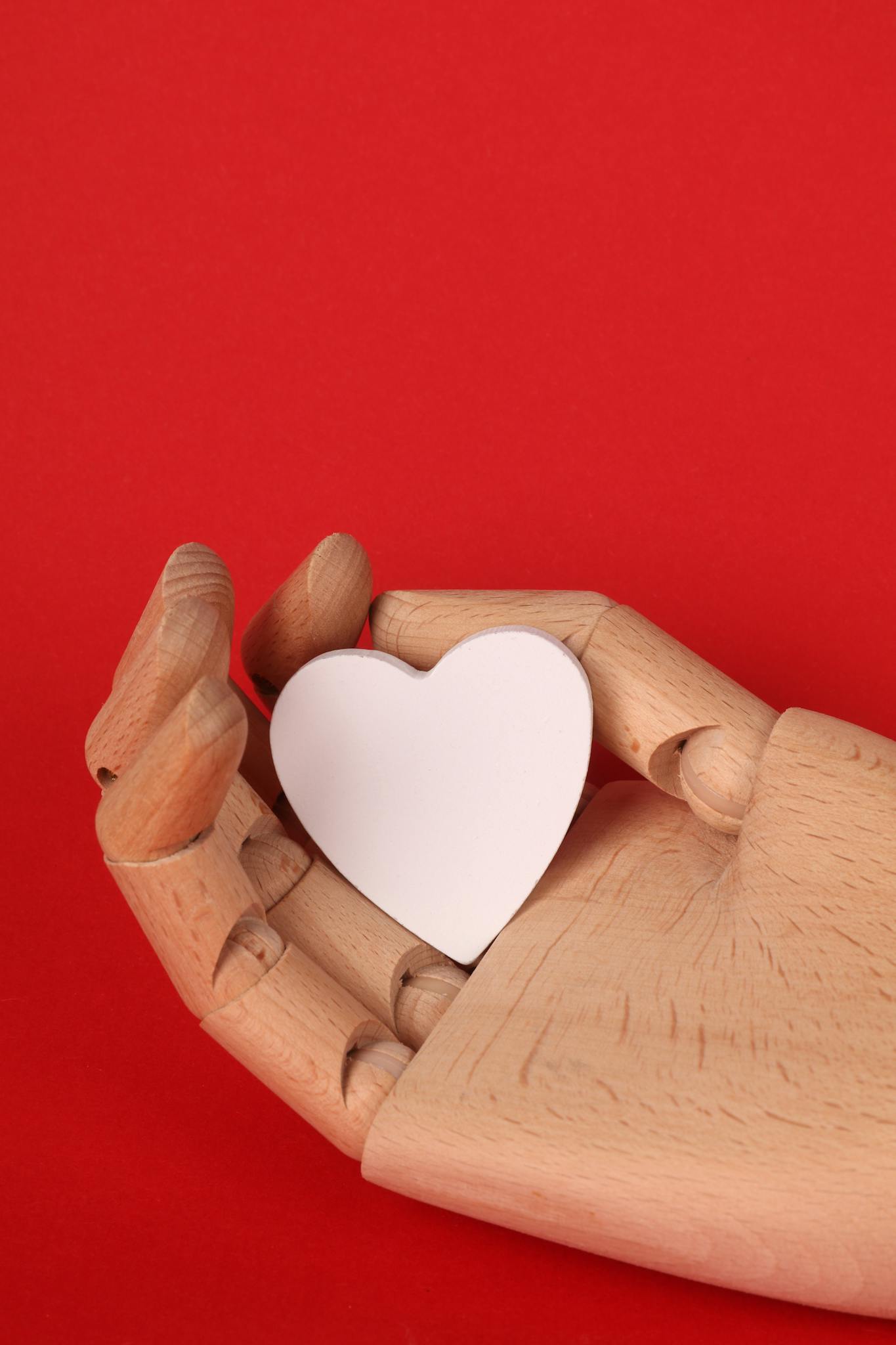 Wooden hand grasping a white heart, symbolizing love and care against a red backdrop.